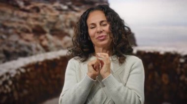 Kıvırcık saçlı ve süveter giyen bir kadın şehir ortamında dışarıda duruyor, sıcak ve güzel bir ifadeyle..