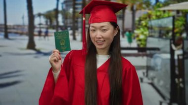 Kırmızı mezuniyet elbisesi giymiş, elinde Çin pasaportu olan deniz kenarında gezinti yapan bir kadın açık havada başarıyı kutluyor..