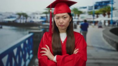 Kırmızı mezuniyet elbisesi giymiş bir kadın deniz kenarındaki gezinti alanında kollarını kavuşturup güneşli bir ortamda ciddi bir ifade takınıyor..