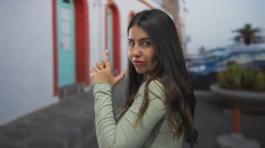 Yeşil tişörtlü genç İspanyol kadın canlı bir sokakta elinde silahla şakacı bir şekilde poz veriyor, dinamik enerji ve kentsel yaşam tarzı görüntüsü sergiliyor..