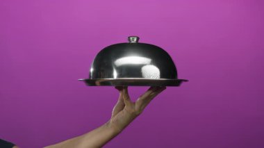 Gümüş servis tepsisini kalın pembe duvara dayayan adam, minimalist izole bir ortamda zarafet sergiliyor, elleri ve metalik yüzeyi vurguluyor..