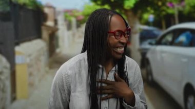 Kırmızı gözlüklü ve örgülü gülümseyen bir kadın, arka planda arabaları olan bir şehir sokağında neşeli bir şekilde el kol hareketi yapar, açık havada neşeli ve çekici bir anı yakalar..