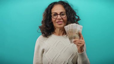 Orta yaşlı bir kadın elinde Rus rublesi banknotları tutuyor, mavi arka planda gözlük ve süveter takıyor, finans ve para birimi temalarını sergiliyor..