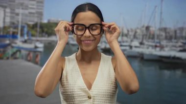 Limanda arka planda teknelerle gülümseyen, gözlüklü ve beyaz bluzlu bir kadın, deniz kenarında zarif bir şekilde neşeli bir atmosfer gösteriyor..