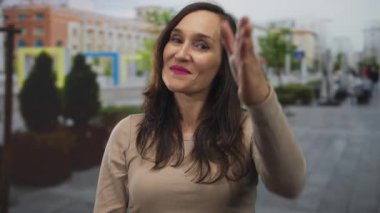 Şehir caddesinde güvenle gülümseyen bir kadın, canlı ruj ve günlük kıyafetlerle elini uzatarak, dost canlısı, kentsel bir atmosfer yaratıyor..