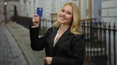 Şehir sokaklarında siyah takım elbiseli, gülen bir kadın elinde mavi bir ödeme kartıyla gün içinde şehir manzarasında neşeli bir ifadeyle gülümsüyor..