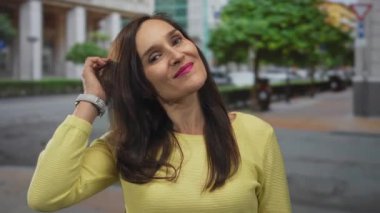 Şehir sokaklarında güvenle gülümseyen bir kadın, sarı bir kazak giyiyor, uzun siyah saçlı ve saatli, şehir çevresi ve yeşilliklerle çevrili..