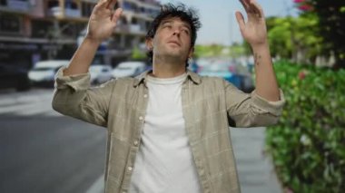 İspanyol bir adam güneşli bir sokakta dikiliyor. Arka planda bulanık arabaları ve yeşilliği var. Açık hava ortamında cazibesini sergiliyor..
