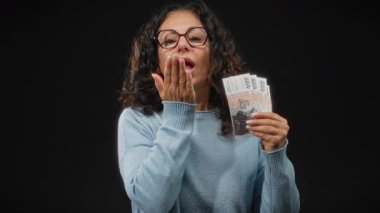 Gözlüklü orta yaşlı bir kadın İzlanda Krona banknotları tutuyor, siyah bir duvara yaslanmış, neşeli bir ifadeyle öpücük yolluyor..