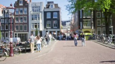 Güneşli bir günde bisiklet ve ikonik Hollandalı binalarla dolu büyüleyici bir kanal caddesinde yürüyen insanları gösteren bulanık bir manzara.
