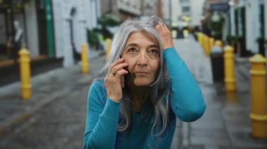 Canlı bir şehir sokağında telefonda konuşan gri saçlı yaşlı bir kadın. Sarı cüsseli ve şehir mimarisinin arka planında..