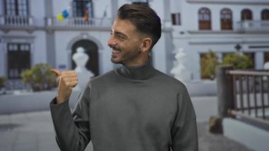 Gülen adam, beyaz bina balkonunun önündeki güneşli şehir caddesini işaret ediyor; dostça bir davet.