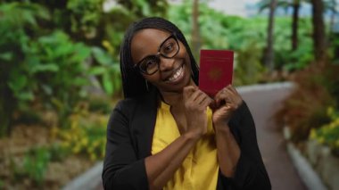 Açık hava parkında Fransız pasaportu taşıyan, gözlüklü ve sarı tişörtlü arka planda yeşillikle gülümseyen seyahat ve çeşitliliği somutlaştıran bir kadın..