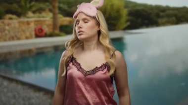 Gecelikli sarı saçlı bir kadın otel havuzunun yanında serinliyor, pembe bir uyku maskesi takarken sıcaklığını ifade ediyor..