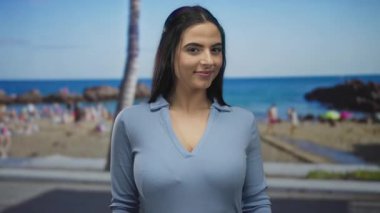 Mavi gömlekli bir kadın okyanus ve palmiye ağacıyla güneşli plaj gününün tadını çıkarıyor. Açık hava rahatlığı ve deniz manzarasının altını çiziyor.