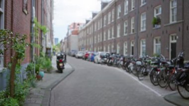 Tuğla binalar boyunca dizilmiş bisiklet ve kaldırım taşlı bir yolda park etmiş scooter ile sakin bir şehir manzarası yaratıyor..