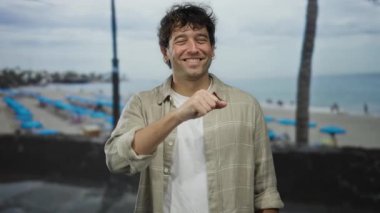 Arka planda palmiye ağaçları ve mavi şemsiyeler olan tropik bir plajda yumruğunu uzatarak kameraya güvenle gülümseyen İspanyol bir adam..