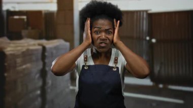 İnşaat alanında önlük giyen bir kadın stresli görünüyor. Kafasını kapalı bir endüstriyel ortamda tutuyor. Bu da işle ilgili bir hayal kırıklığı ya da kaygı olduğunu gösteriyor..