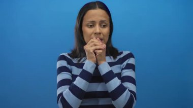 Çizgili kazak giyen bir kadın, mavi bir arka plan üzerinde düşünceli bir şekilde duruyor, elleri birbirine kenetlenmiş duygularını ifade ediyor..