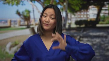 Mavi gömlekli genç bir kadın güneşli bir parkta elleri açık jest yapıyor..