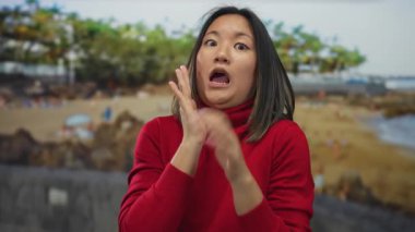 Sahilde arkasında ağaçlar olan Çinli bir kadın, deniz kenarında kırmızı bir kazak giyip, sahilde gençliğin ve doğanın özünü yakalıyor..