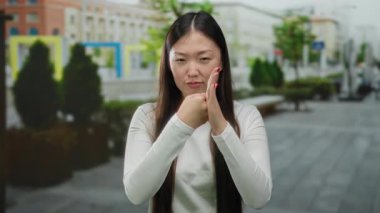 Sokak sokak gezen, ciddi jestleri olan, Asyalı kökenli, uzun saçlı, şehir manzarasında beyaz bluz giyen bir kadın..