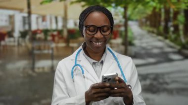 Kadın doktor dışarıda akıllı telefon kullanıyor. Restoran terasında dinleniyor. Güler yüzlü, açık hava ortamında teknoloji ve rahatlama karışımı bir şey sergiliyor..