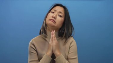 Elleri bağlı dua eden bir kadın mavi bir arkaplan karşısında umudunu dile getiriyor. İçten ve duygusal bir duruş sergiliyor..