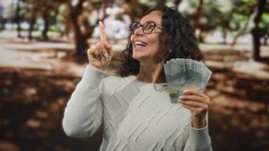 Orta yaşlı bir kadın elinde Endonezya rupiası banknotları tutarken şehir caddesinde durup finansal bir fırsat ya da fikri işaret ediyor..