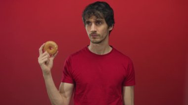 Kırmızı bir stüdyo duvarına karşı düşünceli bir duruşta, jöleli donut tutan genç adam ve çene üzerinde el; merak.