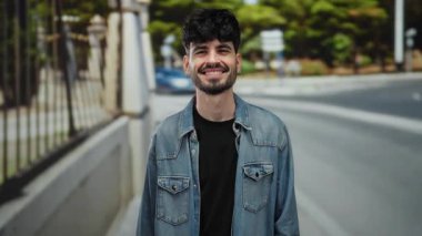 Şehirdeki bir sokakta gülümseyen genç bir adam. Sıradan bir kot ceket giyiyor. Kendinden emin ve rahat görünüyor..