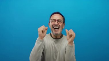 Olgun İspanyol adam coşkulu ve hareketli bir ifade sergileyen canlı mavi bir arka plan karşısında coşkulu ve heyecanlı ellerini kaldırarak sevinç ve heyecan ifade ediyor..