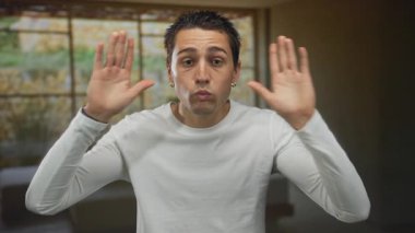 Beyaz uzun kollu gömlek giyen İspanyol genç adam sıcak ışık altında bir otel odasında elleri ile gözlerini kapatır; sürpriz.