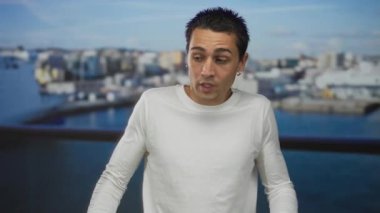 Beyaz elbiseli genç İspanyol bir adam deniz kıyısındaki bir limanda, arkasında şehir manzarası olan teknelerin yanında duruyor, deniz kenarında sakin bir açık hava sahnesi yakalıyor..