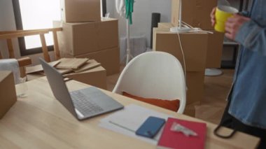 Kot ceketli genç adam, kutularla ve kişisel eşyalarla dolu modern bir evde dizüstü bilgisayarda çalışıyor..