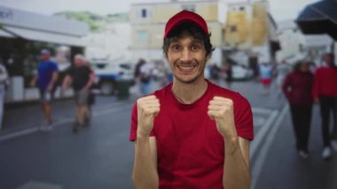 Kırmızı tişörtlü ve şapkalı genç teslimatçı güneşli şehir caddesinde yumruk kaldırıyor ve gülümsüyor; heyecan.