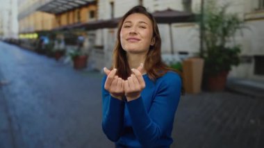 Genç bir kadın şehir sokaklarında gülümserken parmaklarıyla kalp hareketi yapıyor, şehir ortamında mutluluğu ve bağlantıyı ifade ediyor..