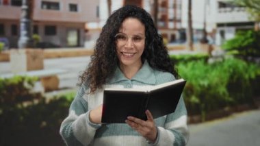 Sokakta siyah defter okuyan genç bir kadın. Arka planda binalar ve yeşillikler, kıvırcık saçlar ve gözlüklerle gülümsüyor..