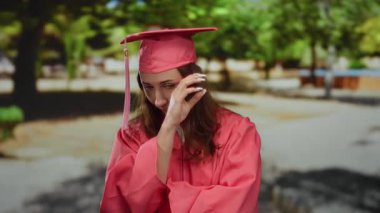 Pembe mezuniyet elbisesi giymiş genç bir kadın güneşli bir parkta, akademik başarı anında duygusal ve huzurlu görünüyor..
