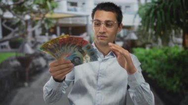 Kentsel caddede elinde Avustralya parası olan genç bir adam, çeşitli banknotlar sergiliyor ve açık havada hareketli bir şehirde gözlük takıyor..