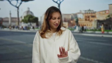 Beyaz kazaklı, belirsiz bir ifadeyle genç bir kadın, klasik mimari ve hafif ışıklandırma ile çevrili bir şehir caddesinde duruyor..