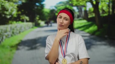 Parkta madalyalı genç bir kadın, kırmızı saç bandı ve beyaz gömlek takıyor, yeşil ve patikayla çevrili, açık havada ilham verici fitness ve başarı hedefleri sergiliyor..