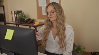 Ofiste, bilgisayar ve sarı saçlı, gözlüklü ve beyaz gömlekli bir kadın, iş yerindeki bitki ve raflarla ilgili yorgunluk ve hayal kırıklığını gösteriyor..