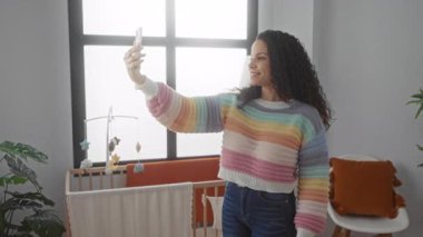 Yatak odasında, pencerenin yanında bebek arabasıyla selfie çeken bir kadın, çağdaş aile yaşamını yansıtan renkli ve sıcak bir ortam izlenimi veriyor..