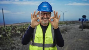 Kasketli ve yelekli olgun bir adam. Yel değirmeni enerjisi arka planında açık hava iş koşullarını gösteriyor..
