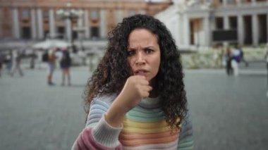 Renkli kazaklı genç İspanyol kadın, arka planda bulanık turistler ve tarihi mimariyle Vatikan 'ın St. Peter Meydanı' nda hayal kırıklığı yaşıyor..