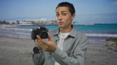 Genç adam deniz kenarında kamera tutuyor, manzaralı sahil manzaralarını yakalıyor, açık hava fotoğrafçılığı becerileri sergiliyor ve canlı kıyı ortamının tadını çıkarıyor..