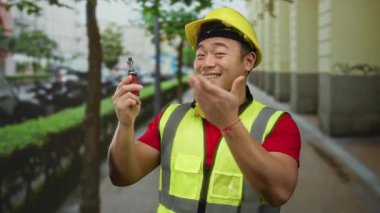 Güvenlik teçhizatı içinde genç Asyalı inşaat işçisi elinde bir ses cihazı tutarken gülümsüyor, modern yaşam tarzını ve şehir ortamını ön plana çıkarıyor..