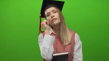 Kitap ve mezuniyet şapkası takmış genç bir kadın yeşil duvara yaslanmış düşünceli ve düşünceli görünüyor.