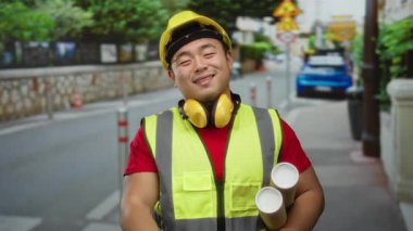 Şehir caddesinde gülümseyen planlı genç bir adam, sarı bir şapka ve yansıtıcı bir yelek giyiyor, kentsel yapıyı somutlaştırıyor..